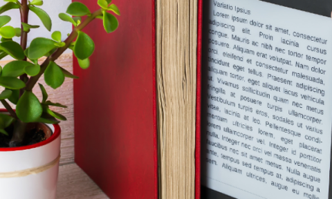 A hand holds an e-reader displaying text between books on a shelf. A small potted plant with green leaves adds a touch of nature to the scene.