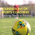 A close-up of a colorful soccer ball on a grassy field with blurred figures in the background. Text reads “Foundations of Sports Coaching, Third Edition.”